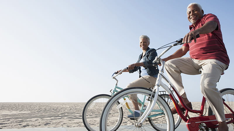 Una pareja de personas mayores de raza negra pasea en bicicleta por una playa.