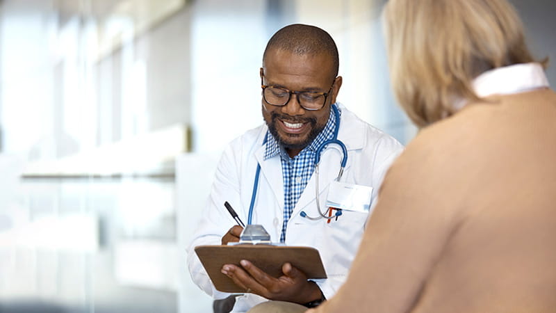 Un médico de raza negra que sonríe toma notas mientras habla con su paciente femenina de edad avanzada.